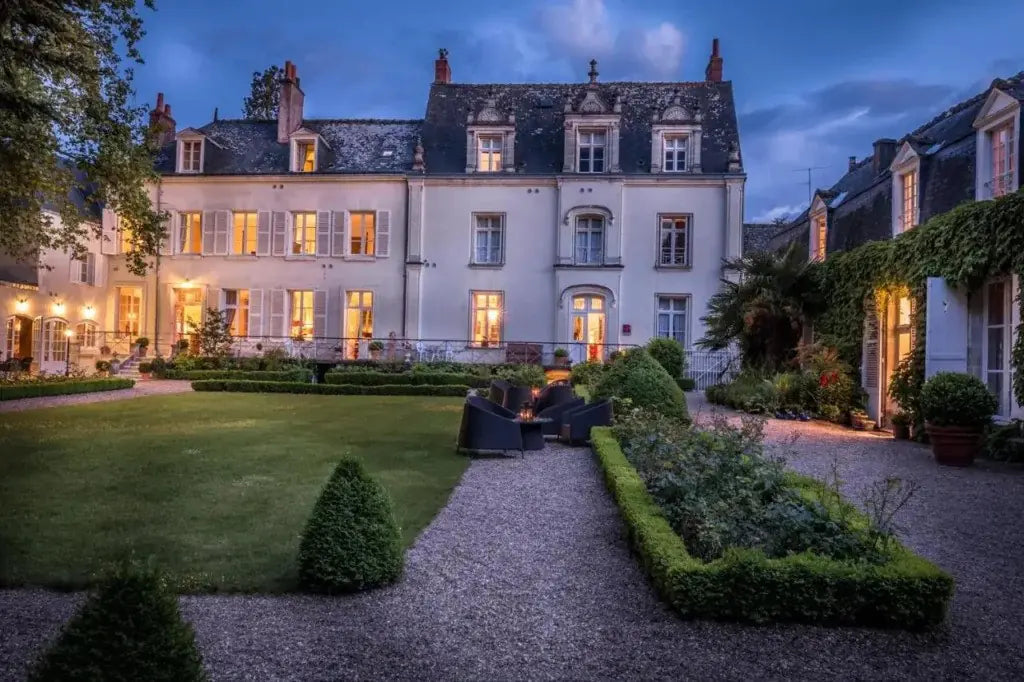 An exterior view of a luxurious hotel in Amboise, France, with lights on and guests seated at a table in front, set against a dusk background.